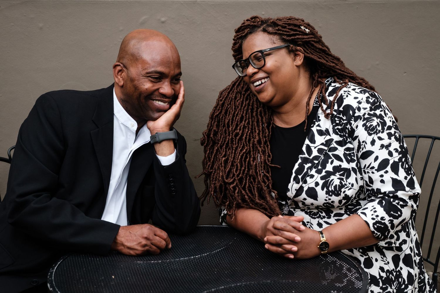 A man in a black suit with a white shirt smiles with a woman wearing glasses wearing a black and white floral print sweater at a table.