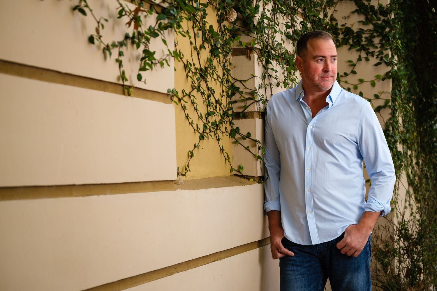 A smiling man wearing a blue shirt and jeans leans against a wall of ivy.