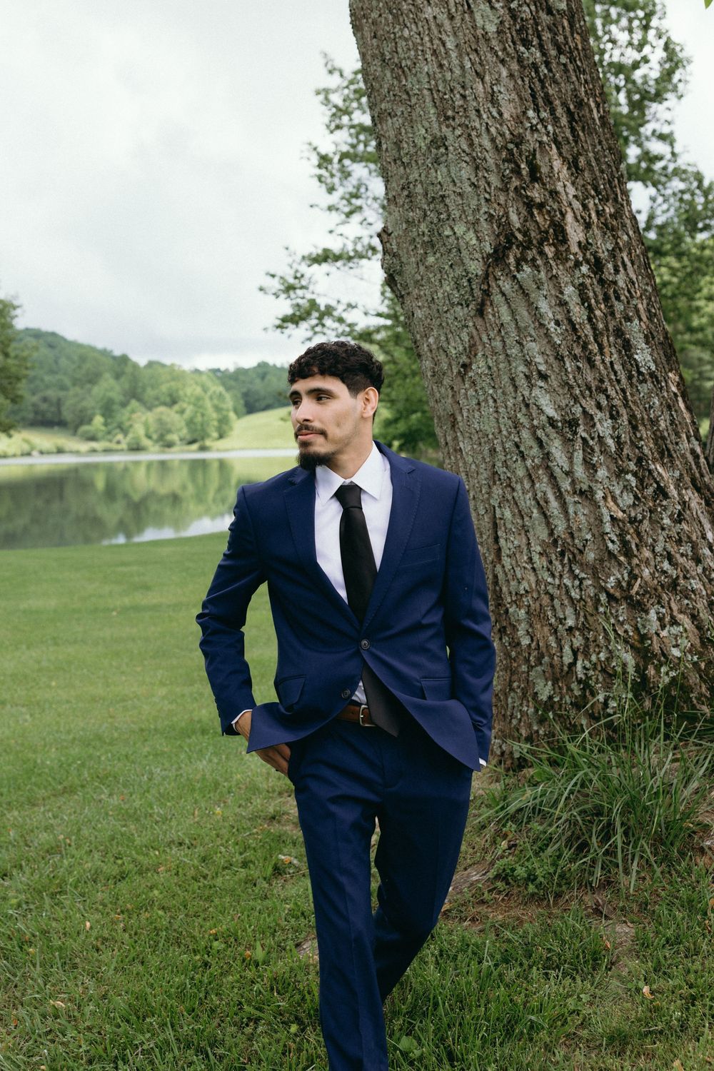 Navy suited figure posed against large tree trunk with lake in background. Estate and historic venue wedding photographer