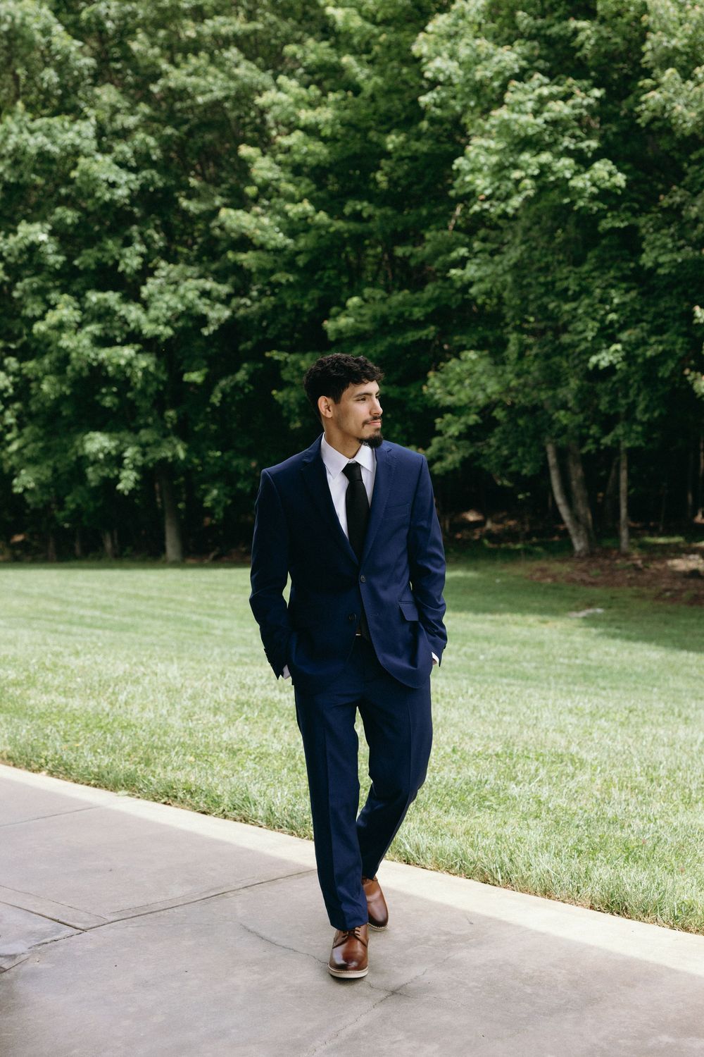 A person in a navy suit walks along a concrete path bordered by lush green grass and trees. Estate and historic venue wedding photographer