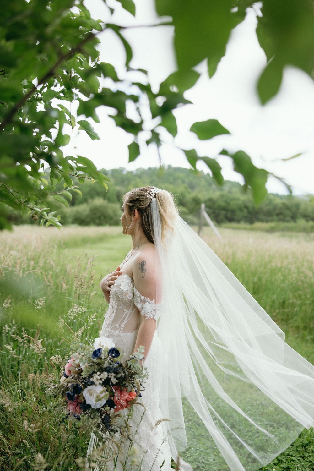 A flowing cathedral veil catches the breeze as bride stands in tall grass with bouquet. Estate and historic venue wedding photographer