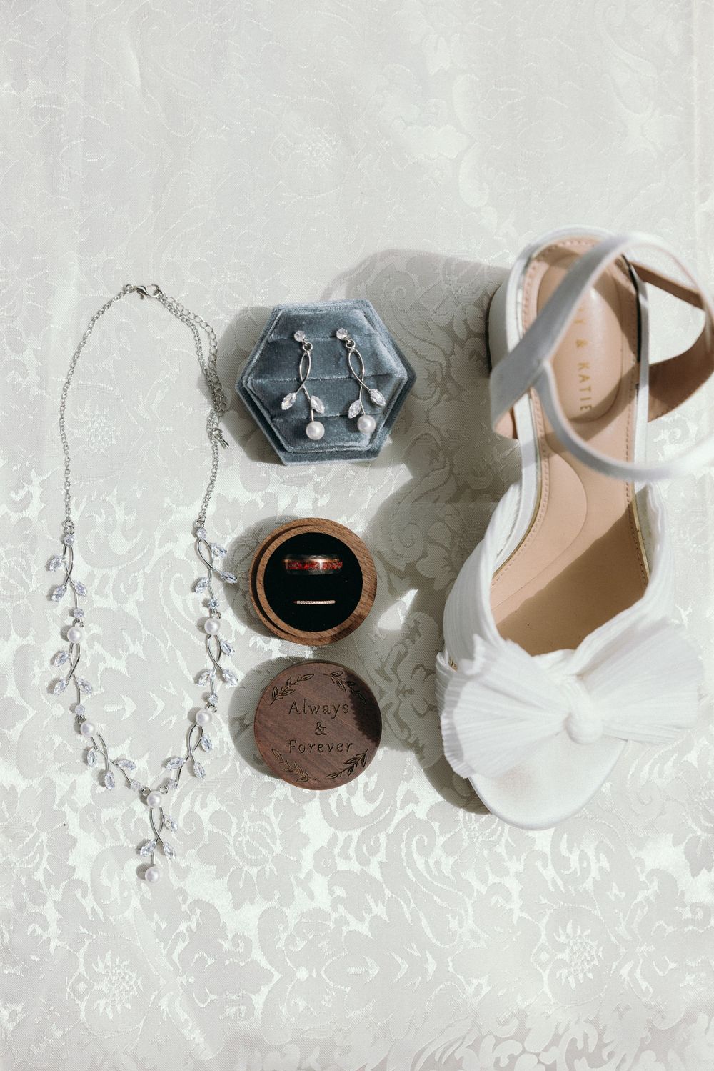 Wedding accessories including white shoes, jewelry, and ring boxes arranged on lace fabric. Estate and historic venue wedding photographer