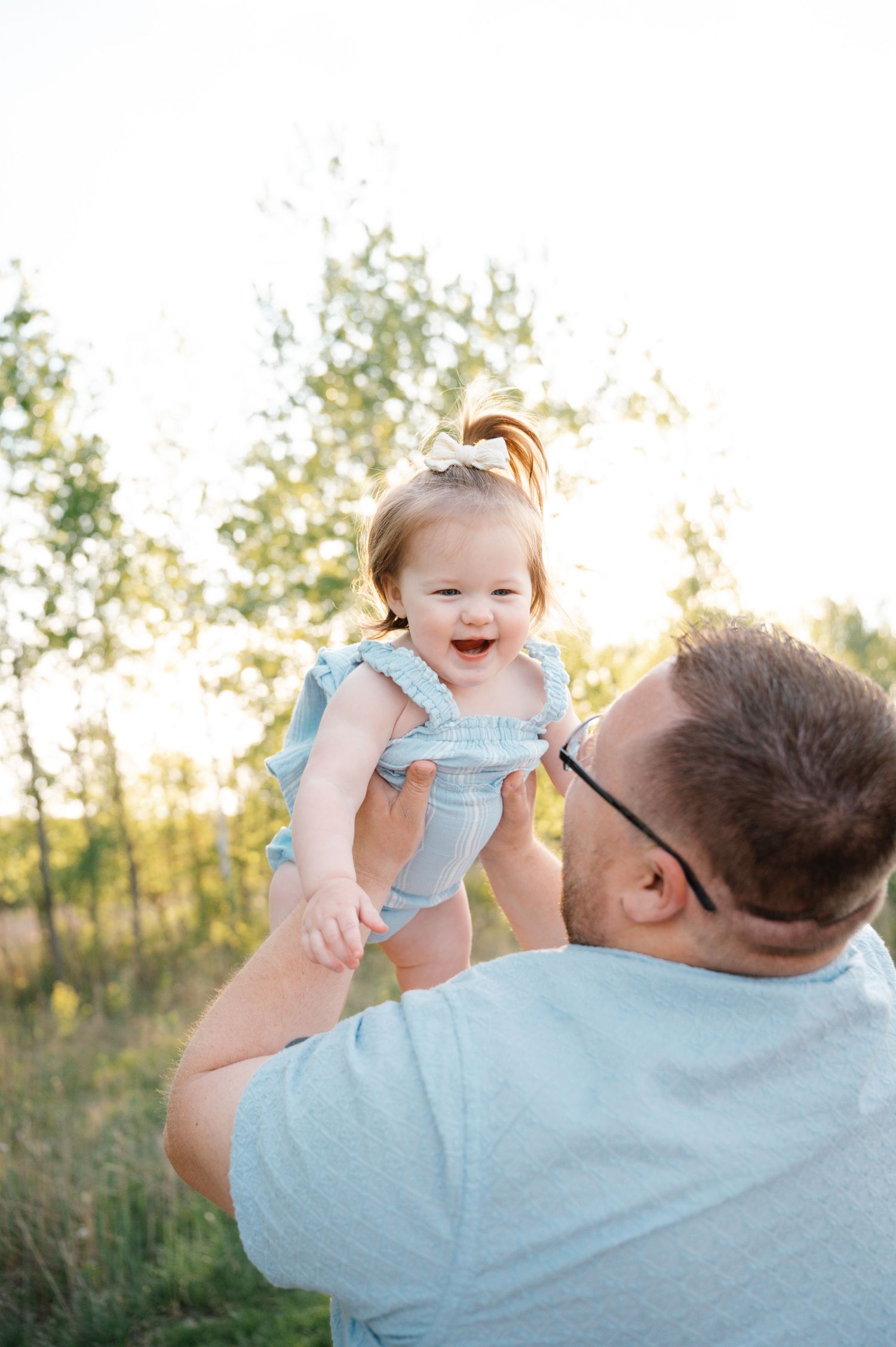 A joyful baby girl in a blue romper laughs as her father lifts her in golden sunlight.