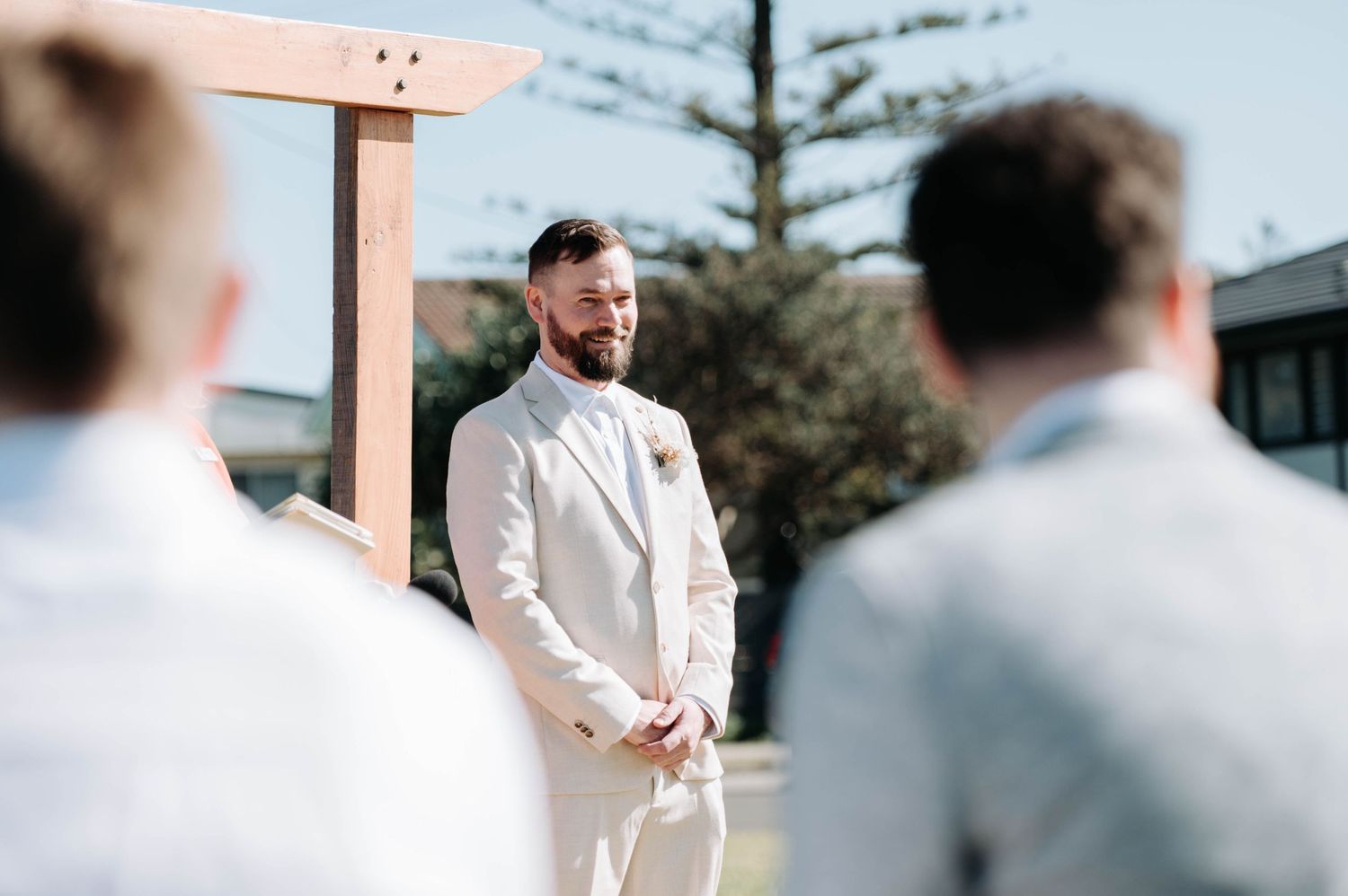 Outdoor wedding ceremony with wooden arch and guests seated on benches under a blue sky.