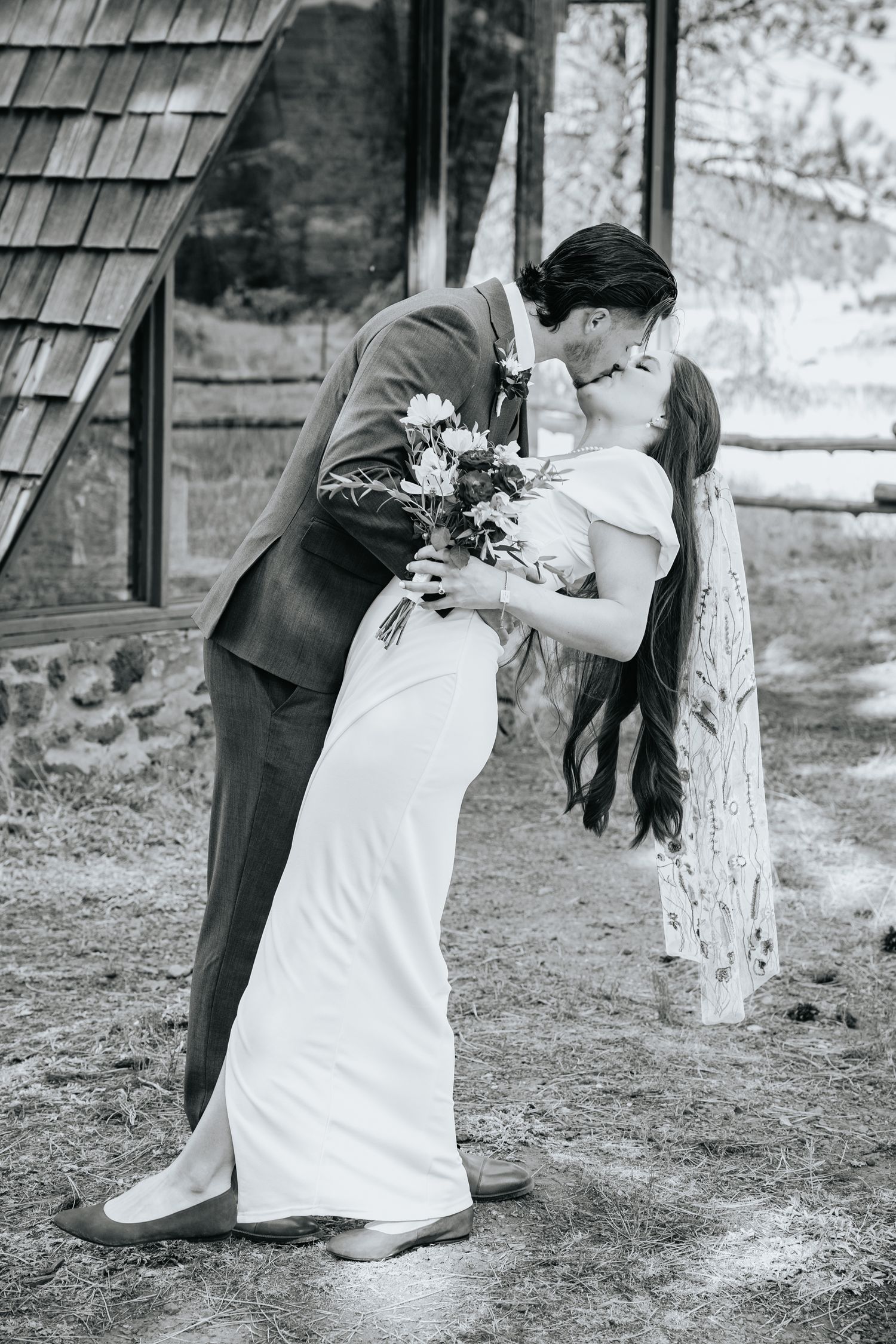 A romantic black and white wedding photo shows a couple sharing a kiss while holding a floral bouquet outside a rustic building.