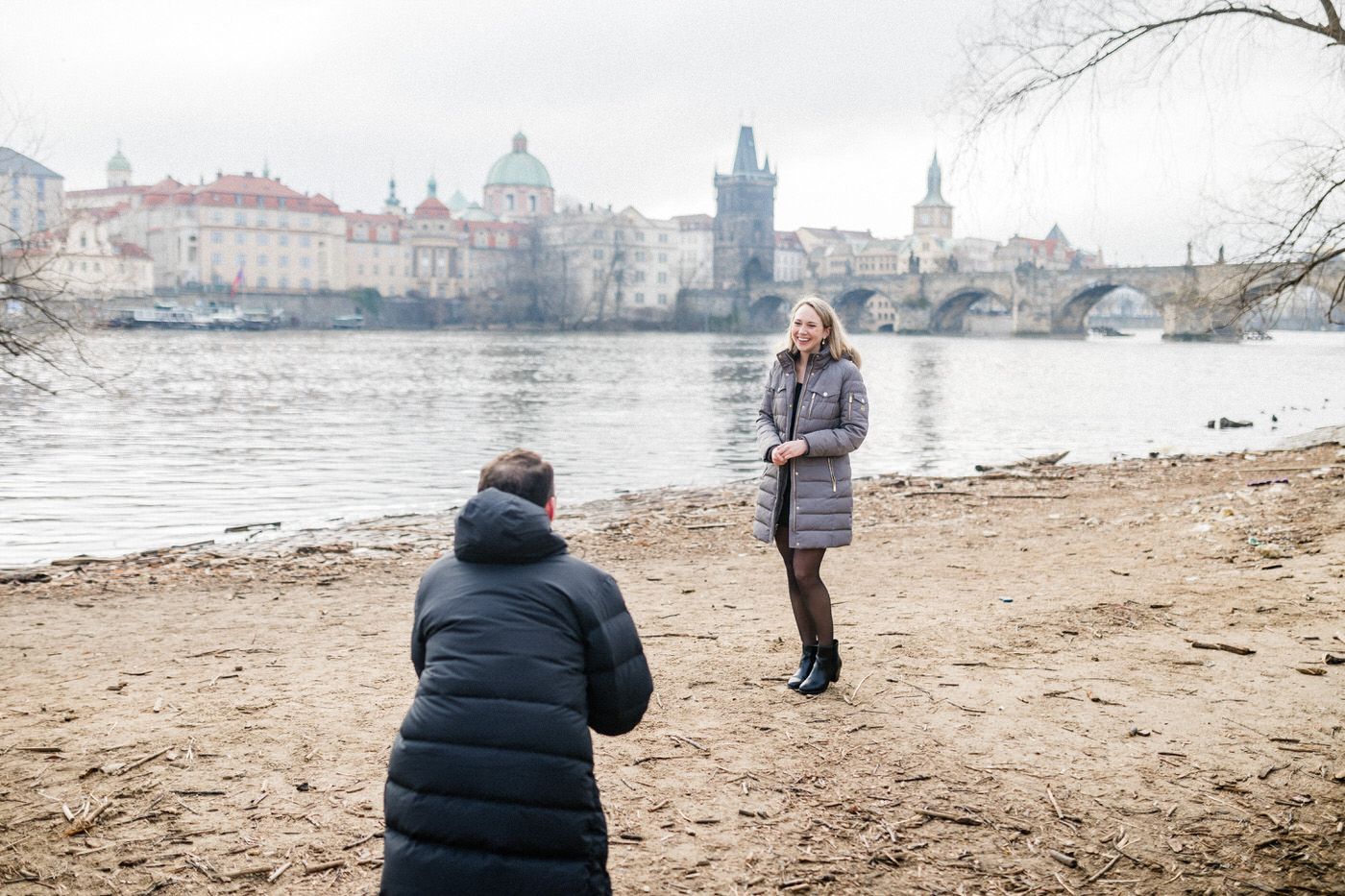 Proposal photography session on a sandy riverbank with historic Prague architecture in the background.