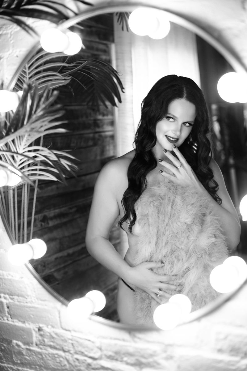 Black and white boudoir portrait with vintage-style lighting, palm plants and vanity mirror bulbs creating a glamorous atmosphere.
