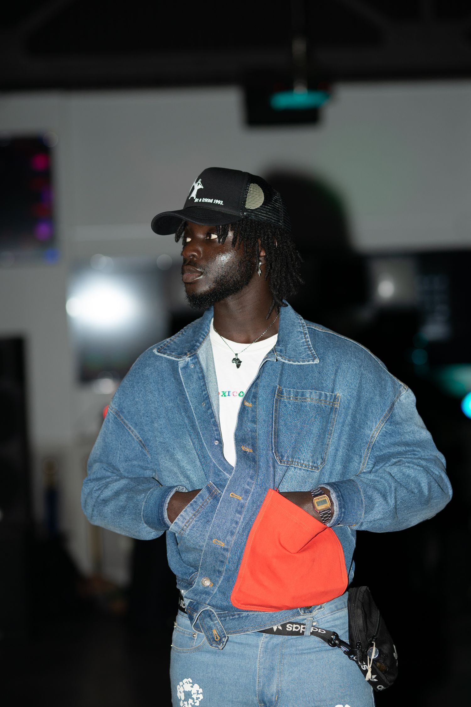 Street style fashion featuring denim jacket, orange accessory and baseball cap in dark setting.