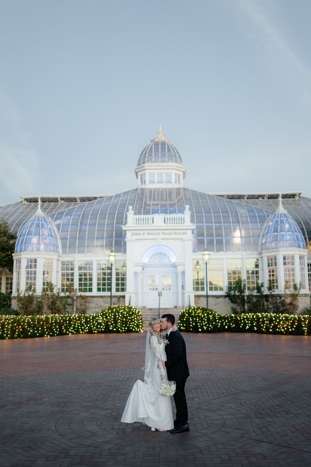 A stunning glass conservatory wedding venue illuminated at dusk with a couple in the foreground.