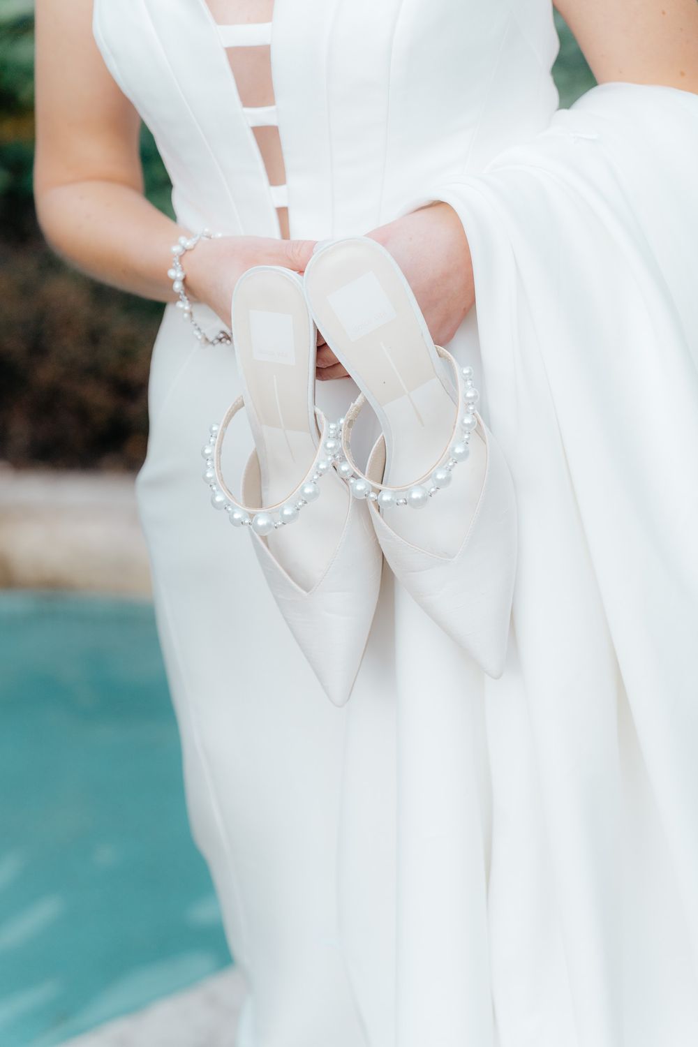 Bride holding elegant white pointed-toe wedding shoes with crystal embellishments.