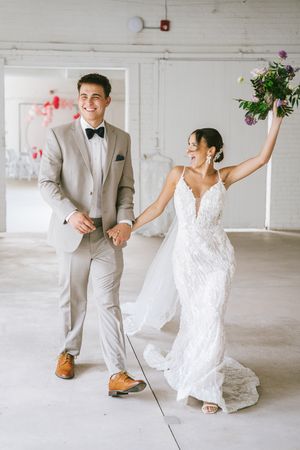 A joyful celebration in a white industrial space with elegant wedding attire and raised bouquet.