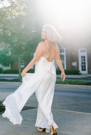 A flowing white bridal jumpsuit twirls gracefully in golden sunlight against a brick building backdrop.