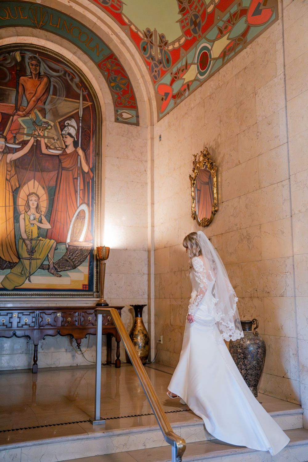 A bride in a white wedding gown stands near an ornate mural and arched doorway.