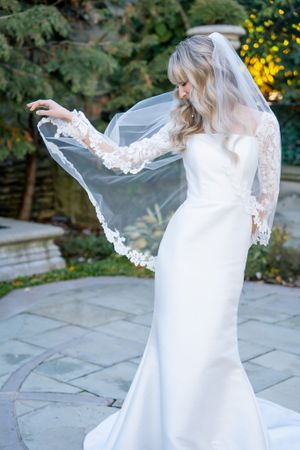 A bride in a long sleeve lace wedding dress playfully holds her veil in a garden setting.