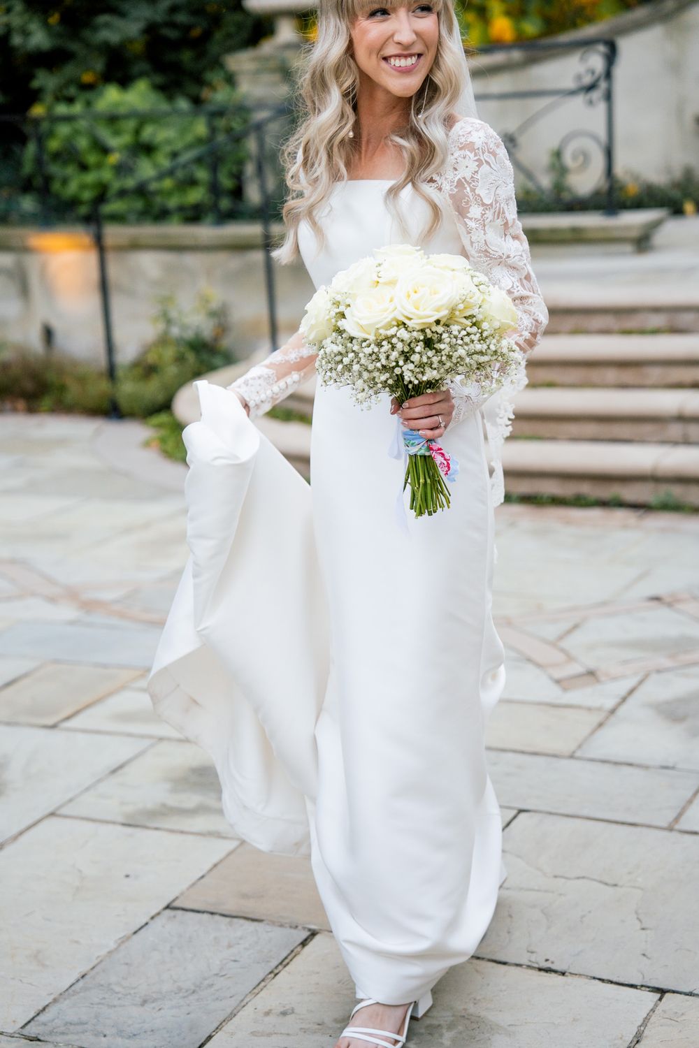 A bride holds a white rose bouquet while wearing an elegant lace-sleeved wedding gown.