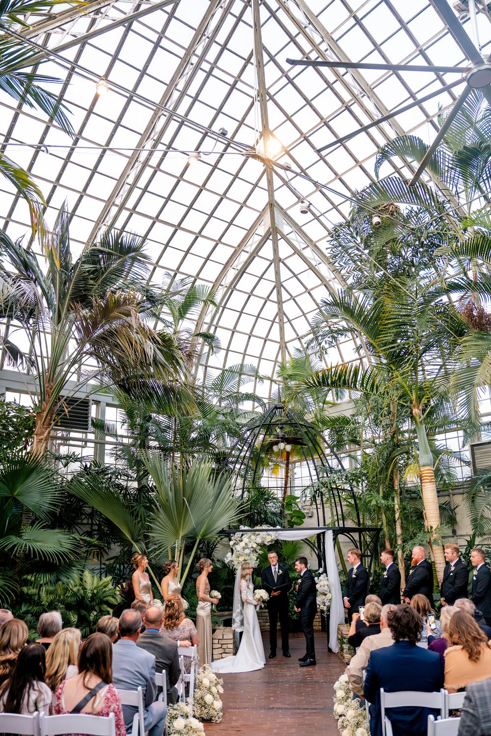 Wedding ceremony taking place in a stunning glass conservatory filled with palm trees and natural light.