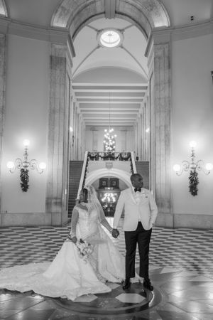 Black and white photo of wedding couple in grand hallway with chandeliers and arched ceiling.