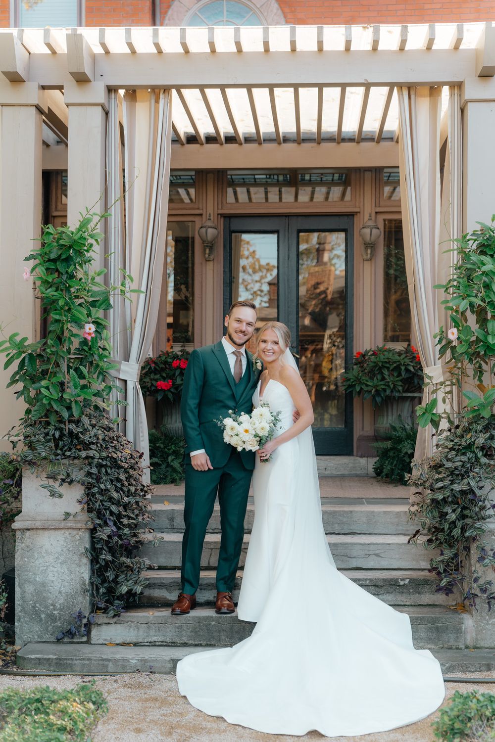 Newlyweds pose under pergola entrance with trailing vines and white curtains.