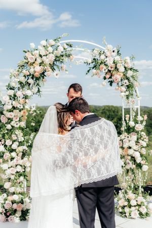A romantic moment under a floral arch decorated with white and pink roses against a blue sky.