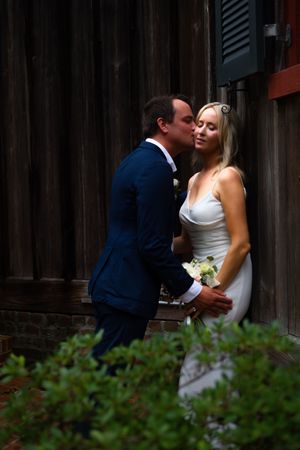 A romantic wedding couple shares intimate kisses against a rustic wooden wall with greenery in the foreground.