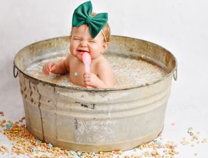 Baby with green bow sits in vintage metal tub surrounded by sparkly confetti.