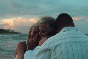 A couple embraces on a beach at sunset with ocean waves and cloudy sky in the background.