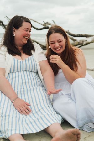 Two friends share joyful laughter while relaxing on a beach with driftwood and overcast skies in the background.
