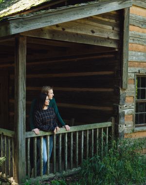 A rustic wooden cabin porch with brick pillars and green grass growing around the weathered structure.