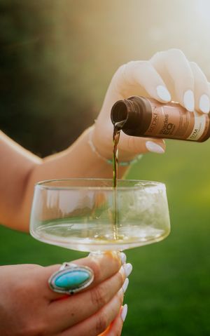 Hand with white nails pours dark liquid from a small bottle into a clear glass against a blurred green background.