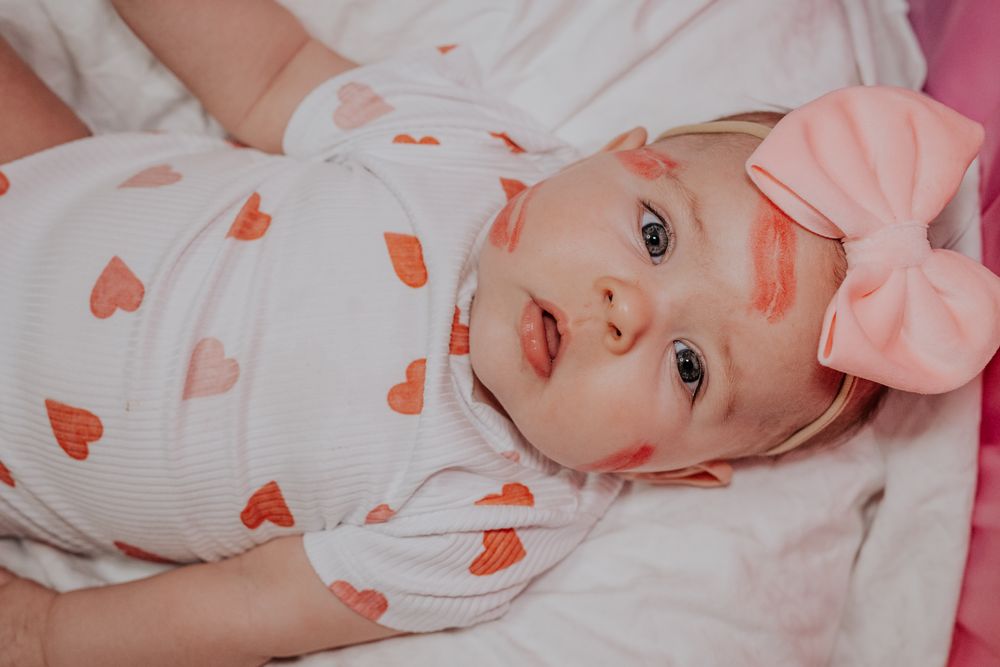 Baby wearing heart-patterned onesie and pink bow lying on white bedding.
