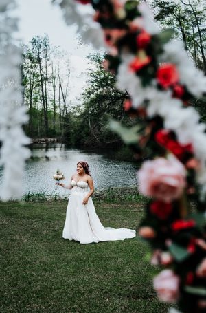 A bride in a white wedding dress stands by a lake surrounded by trees and floral arch decorations.