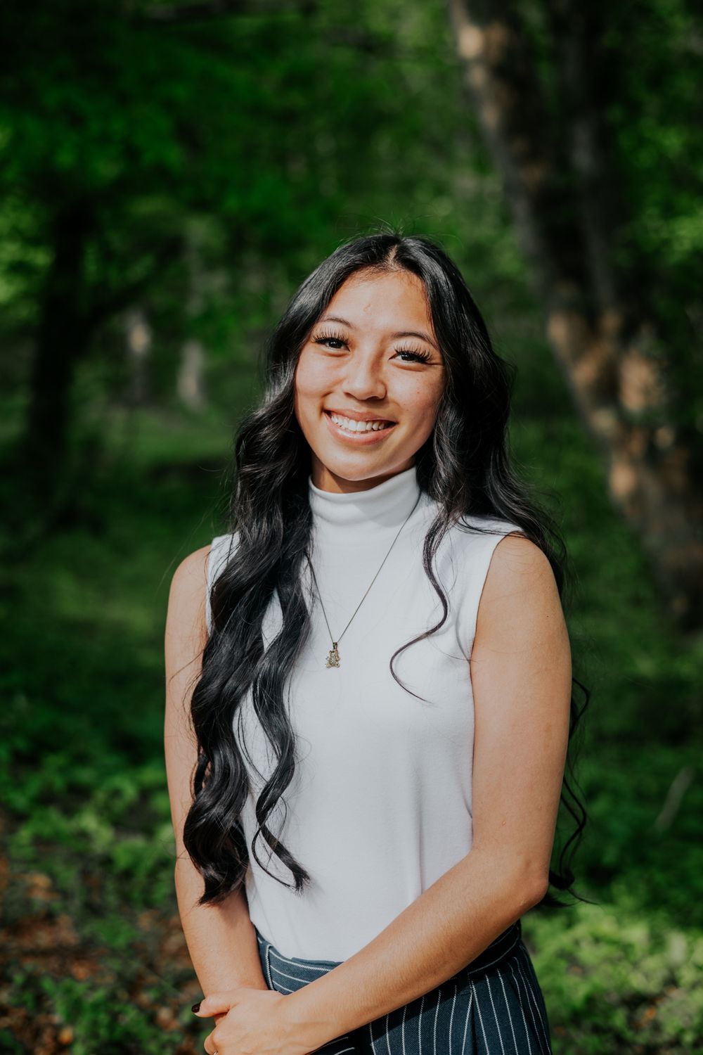 Portrait of person in white turtleneck sleeveless top against lush green forest background.