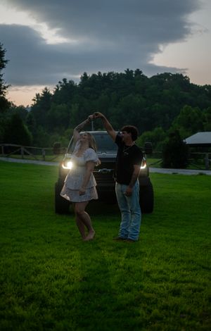 Two figures dance together in a grassy field at dusk with car headlights illuminating the scene.