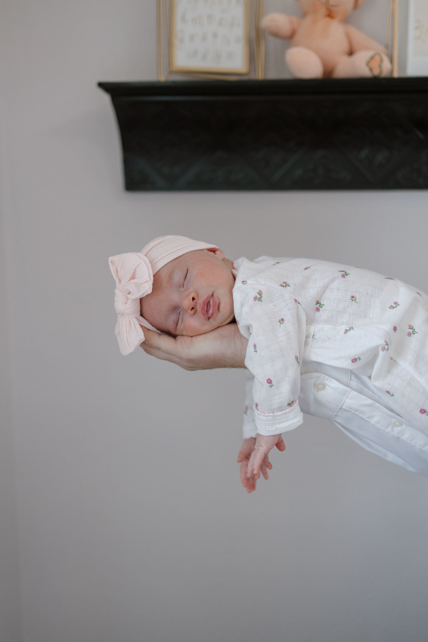 Sleeping newborn baby wearing floral pajamas and pink bow headband resting on white blanket.