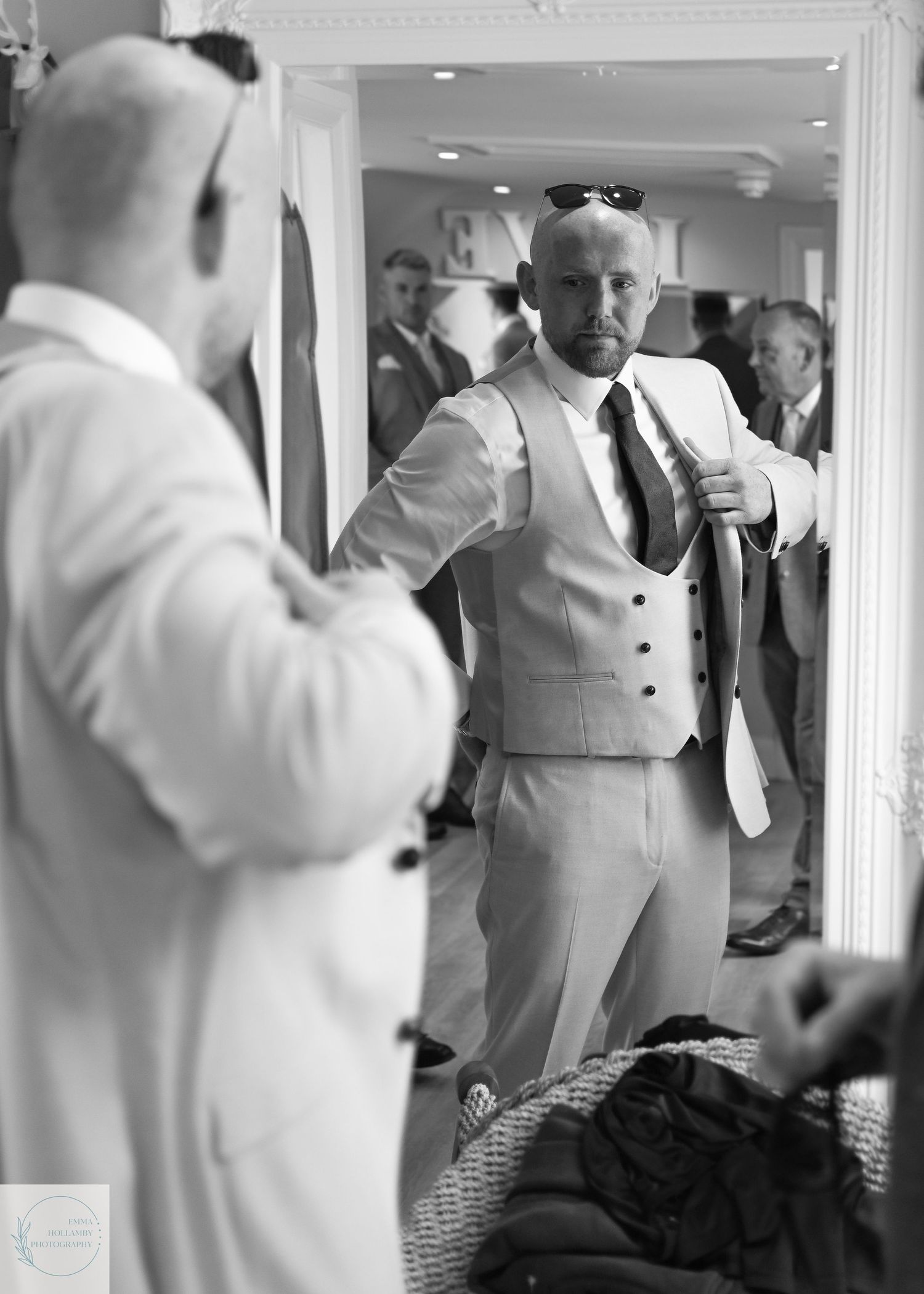 Black and white photo of a bald groom in a light suit adjusting his tie in a mirror.