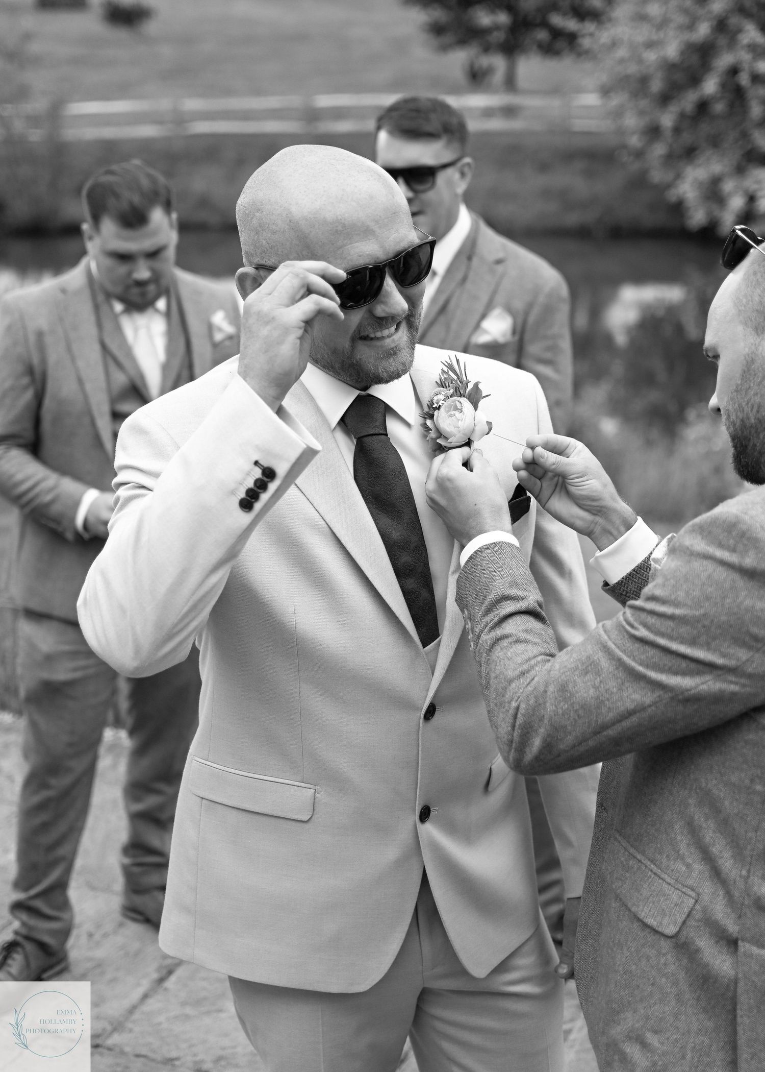 Bald groom in light suit and sunglasses having boutonniere pinned on by groomsman outdoors.