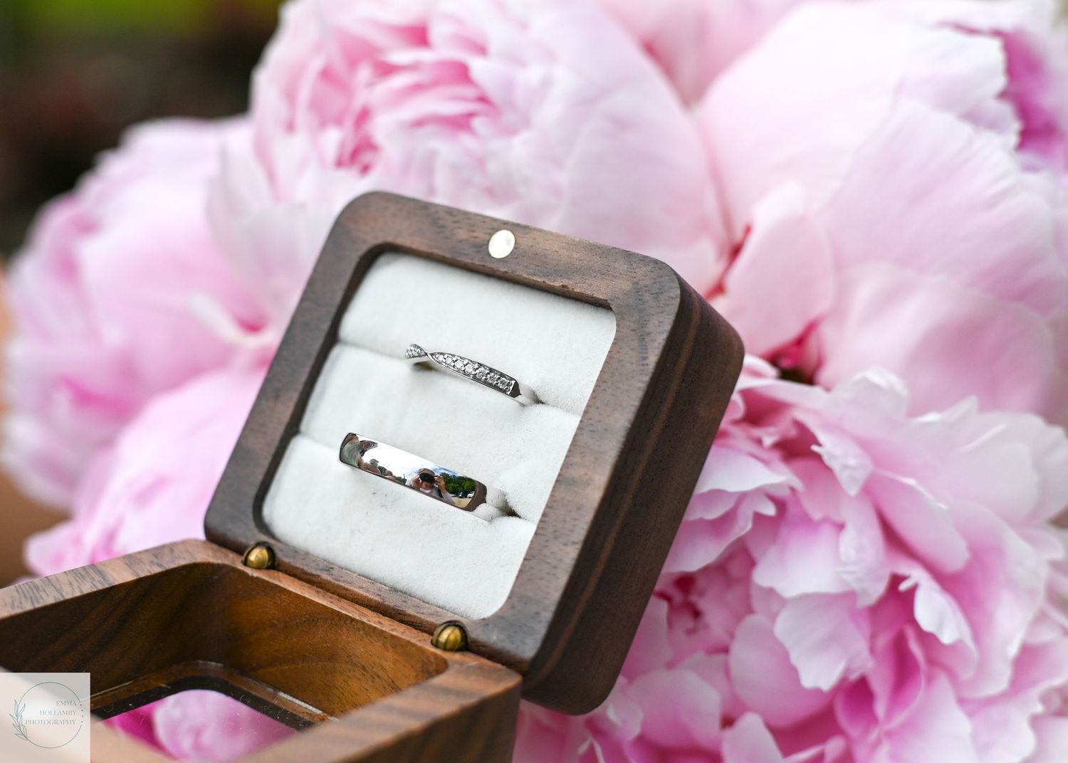 Two wedding rings displayed in a wooden ring box surrounded by pink peony flowers.