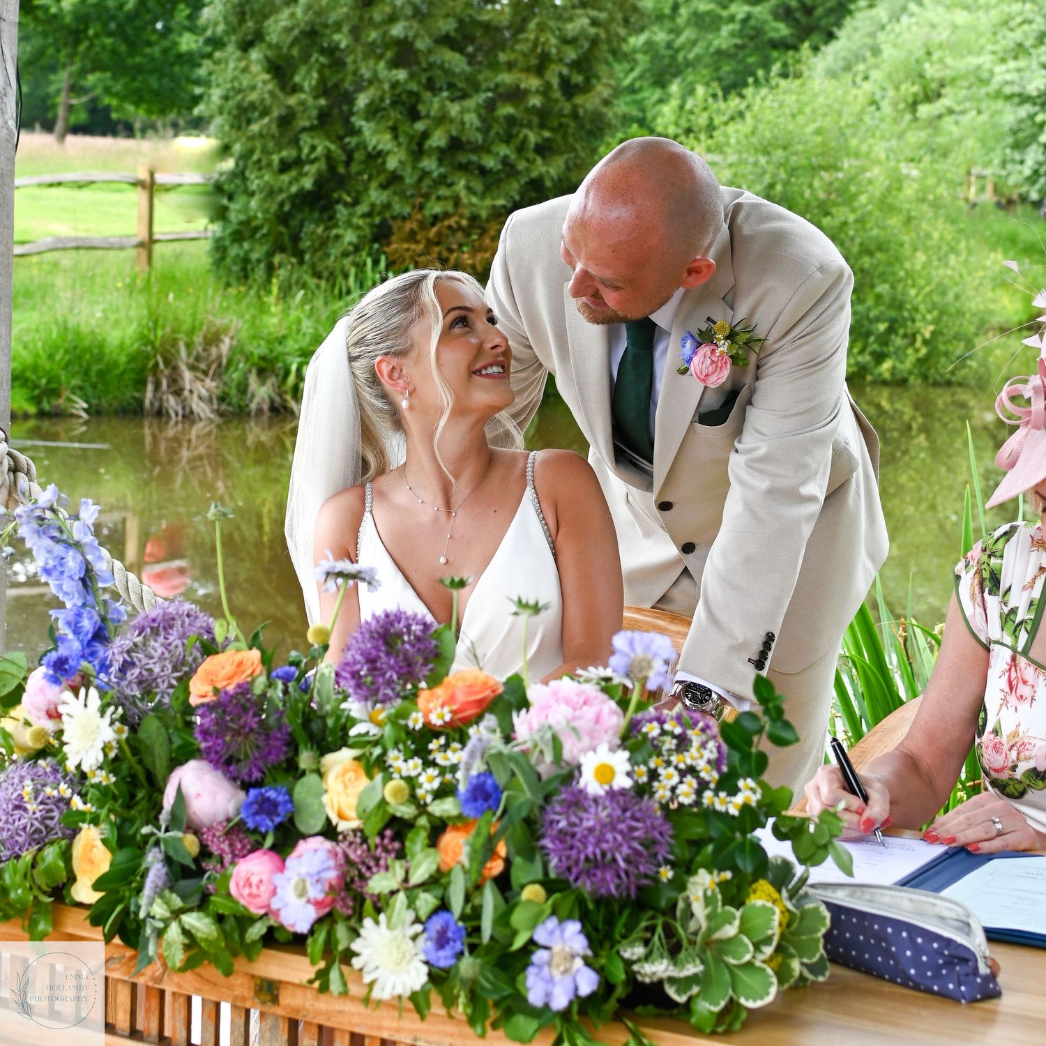 Bride and bald groom share a joyful moment signing the wedding register outdoors by a pond.
