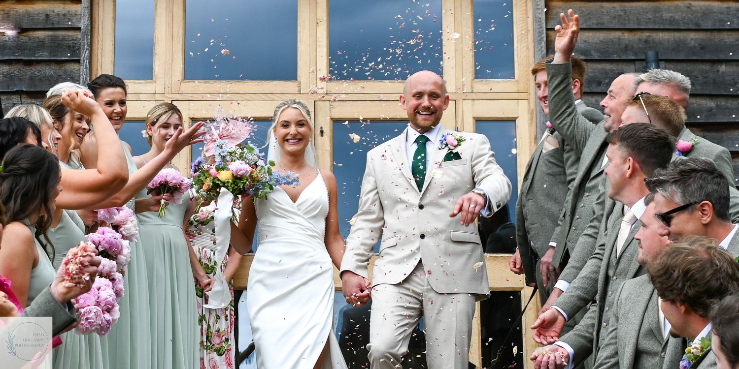 Newlywed couple walks through confetti shower as wedding guests celebrate outside a barn venue.