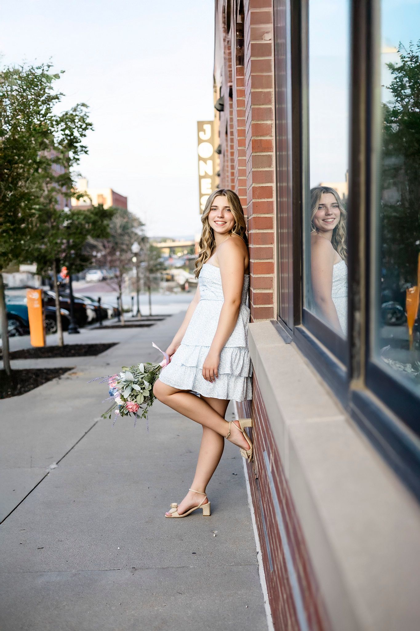 Woman in white dress sits on ledge outside brick building with windows and urban street view in background.