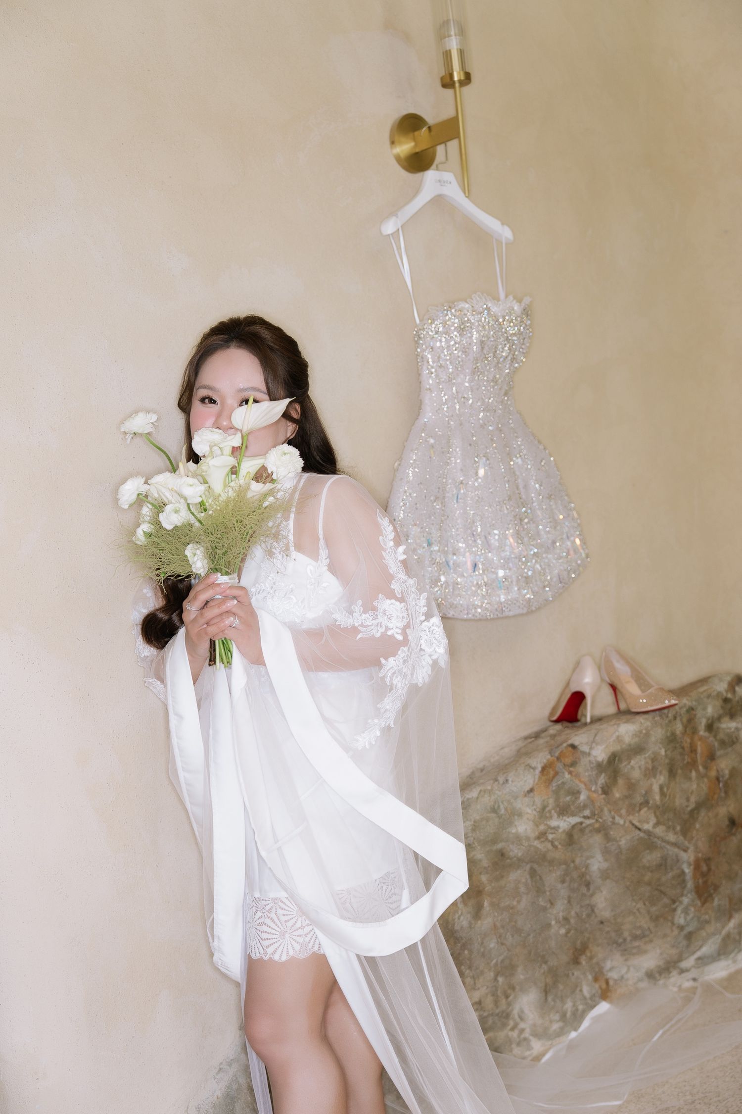 Bride in sheer lace robe holds white floral bouquet beside a sparkling sequin mini wedding dress.