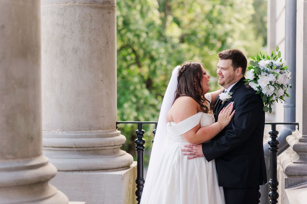 A romantic couple shares a tender moment between elegant stone columns during an outdoor wedding photo session.