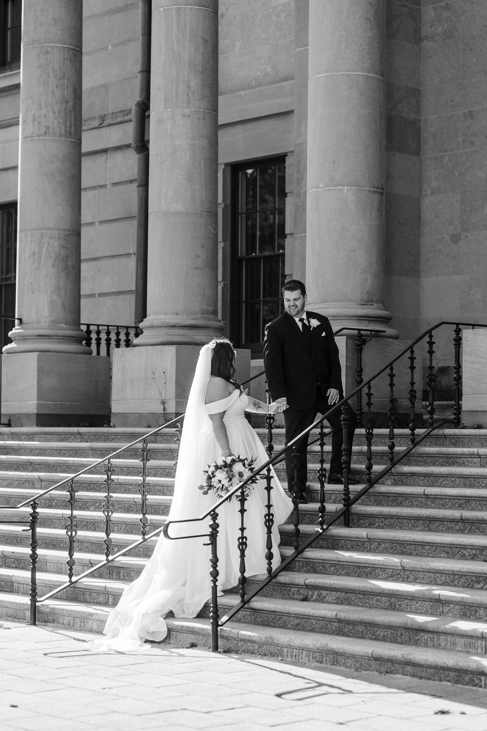 Two figures in wedding attire embrace on elegant stone steps of a columned building in black and white photography.
