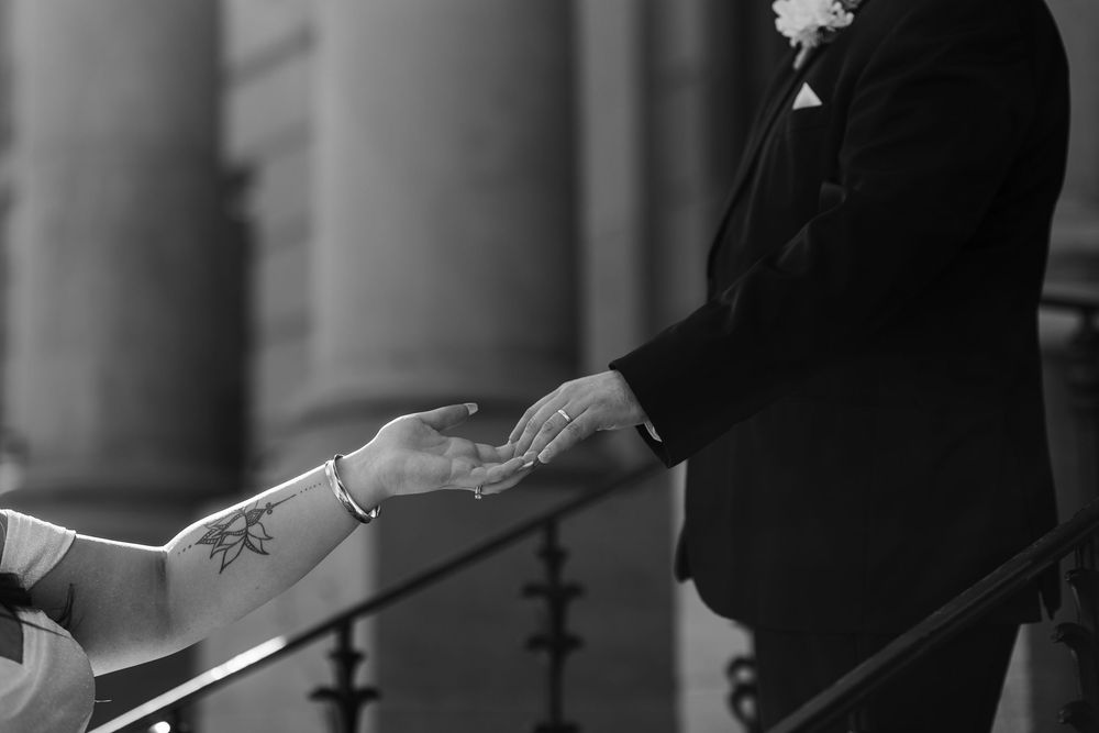 Elegant wedding couple holds hands and shows their wedding rings against a blurred background of pillars.