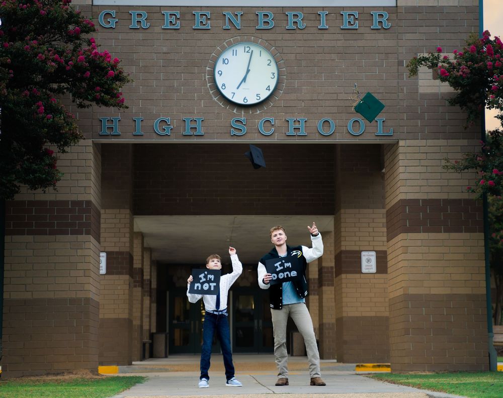 Two high school seniors tossing caps in front of Greenbrier High School entrance with clock above.