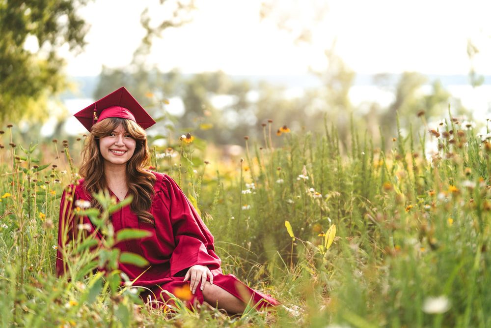 Graduate in red cap and gown sitting in a wildflower meadow with golden sunlight.