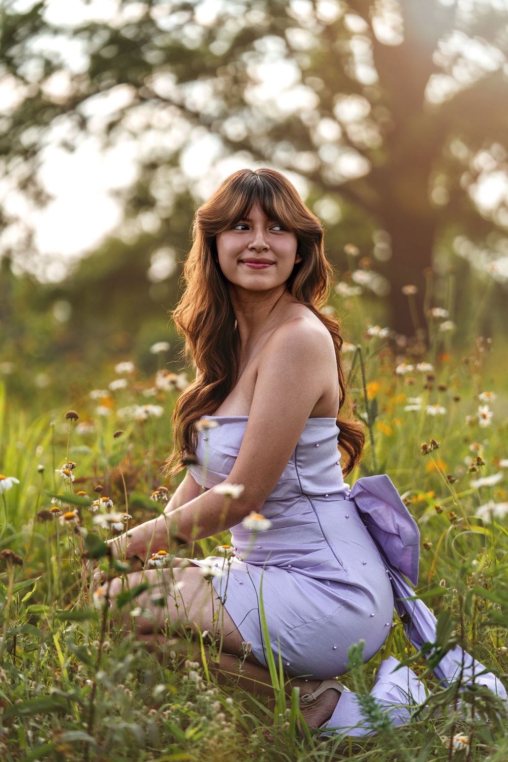 Young woman in lavender dress sitting among wildflowers in a sunlit grassy meadow.