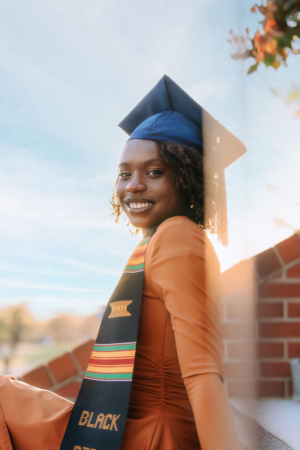 Smiling graduate in blue cap and kente stole with orange dress posing in golden sunlight.