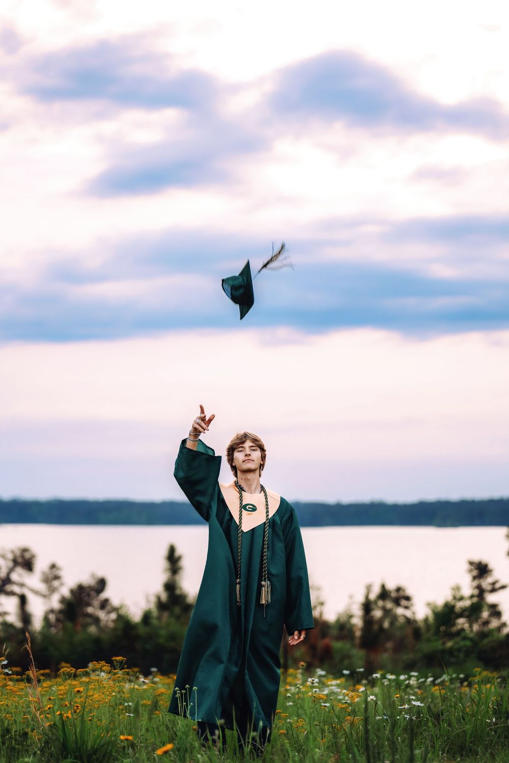 Graduate in green cap and gown tossing mortarboard into cloudy sky near lakeside meadow.
