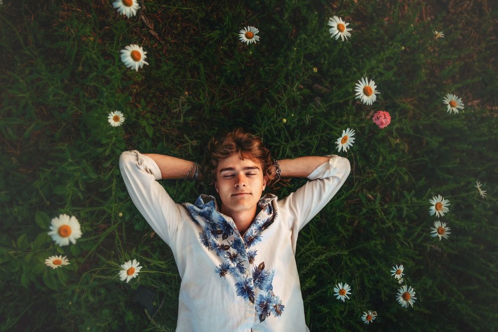 Young person lying in green grass surrounded by white daisies, hands behind head, wearing floral embroidered shirt.